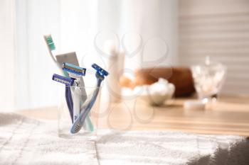Glass with razors, tooth paste and brush on table in bathroom�