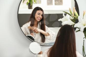 Beautiful young woman combing her healthy long hair near mirror at home 