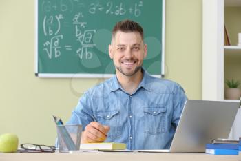 Handsome male teacher with laptop sitting at table in classroom�