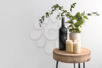 Beautiful burning candles with eucalyptus in vase and bottle of wine on table against light background�
