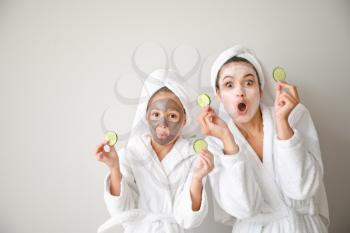 Funny mother and her little daughter with facial masks and cucumber slices on light background 