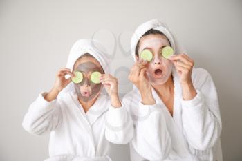 Funny mother and her little daughter with facial masks and cucumber slices on light background�