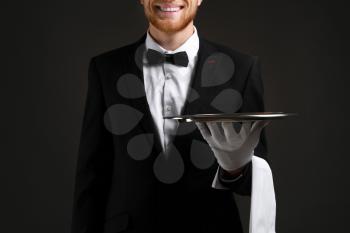 Handsome waiter with empty tray on dark background�