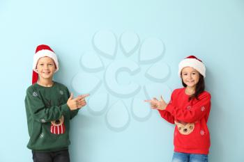 Little children in Christmas sweaters and Santa hats pointing at something on color background�