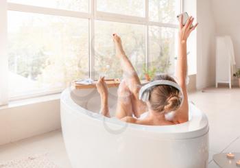 Beautiful young woman listening to music and drinking cocktail while taking bath at home�
