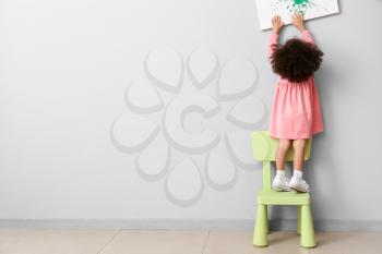 Little African-American girl standing on chair near light wall. Child in danger�