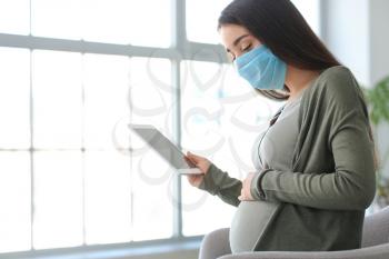 Pregnant woman with tablet computer wearing medical mask at home�