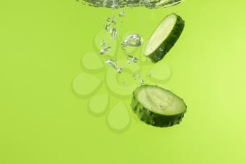 Falling of fresh cucumber slices into water against color background�