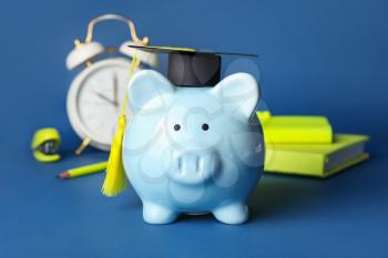 Piggy bank with graduation hat, stationery and alarm clock on color background�