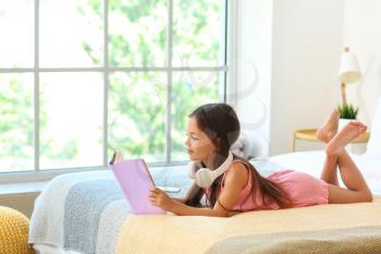 Cute little girl with headphones reading in bedroom�