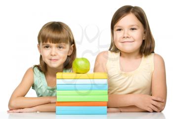 Cheerful little girls with books and green apple are sitting at table, isolated over white