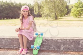 Stylish little girl child with skateboard sits in a park