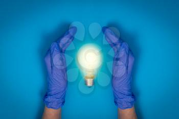 Hands with protective gloves protecting glowing light bulb over a blue background