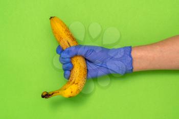 Hand in a latex glove holding a banana on green background with copy space. Grocery stores during COVID-19 lockdown.