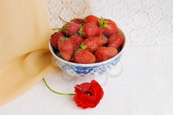Dishes with ripe, red strawberries and red flower on the table with a white cloth on a background of beige and white shades