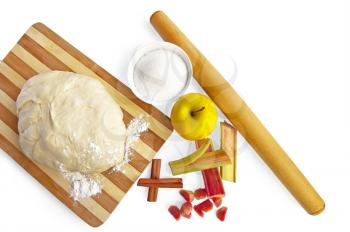 Dough on a wooden board, rolling pin, apple, rhubarb and sugar in a cup, two cinnamon sticks isolated on a white background