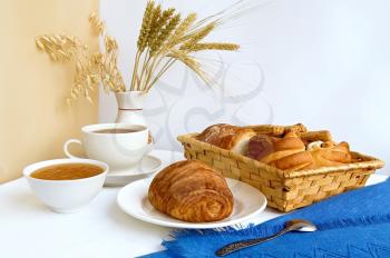Table with a cup of tea and croissant on a plate, rolls in a wicker basket, jam in a bowl, stalks of wheat, oats and rye in a vase, blue cloth, a teaspoon against a background of white and beige curta