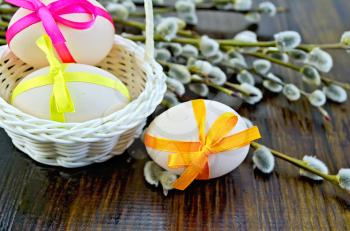 Three Easter eggs tied with colored ribbons, white wicker basket, willow twigs on a wooden board