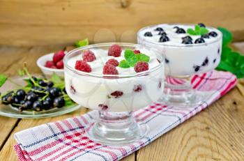 Thick yogurt in two glasses with raspberry and black currant on a napkin, berries and mint on a wooden boards background