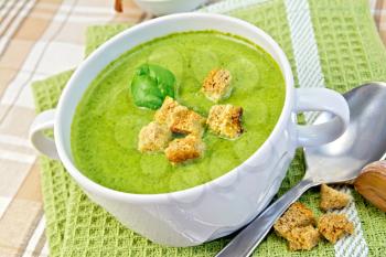Green soup puree in a white bowl with croutons and spinach leaves, spoon, garlic on a napkin on the background fabric
