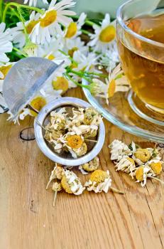 Herbal tea in a glass cup, metal sieve with dry chamomile flowers, fresh flowers, daisies, doily on a wooden board