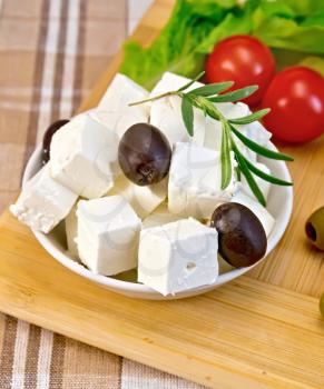 Feta cheese, olives, rosemary in a white bowl, tomatoes, green salad on the background of wooden boards and brown tablecloth