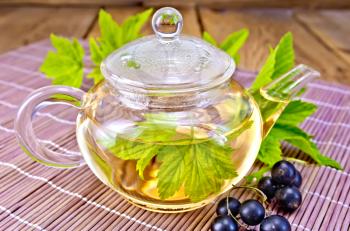 Tea in a glass teapot with leaves of black currant, berries and green leaves of a black currant on a bamboo napkin on a wooden boards background