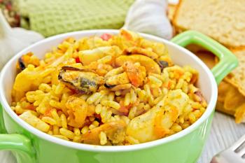 Rice with seafood and saffron in a green pot, napkin, fork, garlic bread on a wooden boards background