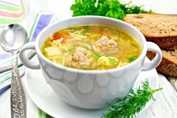 Soup with meatballs, noodles and vegetables in a white bowl, bread, napkin and spoon on the background light wooden boards