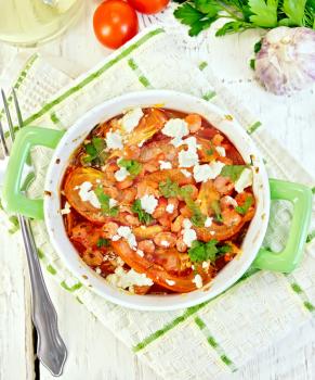 Shrimp and tomatoes baked with feta cheese in a roasting pan on a towel, parsley and garlic on a wooden boards background on top