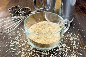 Sesame flour in a glass cup with strainer, mixer and molds for cookies, sesame seeds on background of dark wood planks