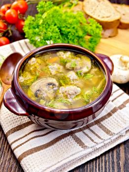 Soup with meatballs, noodles and champignon in a clay bowl on a napkin, parsley, tomatoes, mushrooms and bread on dark background wooden plank
