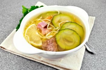 Soup with zucchini, beef, ham, lemon and noodles in a bowl on a napkin, parsley and dill on a granite table background