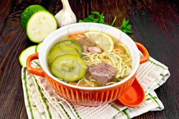 Soup with zucchini, beef, ham, lemon and noodles in a bowl, parsley and dill on a towel on a wooden board background
