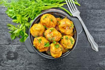 Meatballs with tomato sauce in a brazier with parsley, dill, fork on a black wooden board background on top