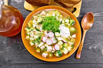 Cold soup okroshka from sausage, potatoes, eggs, radish, cucumber, greens and kvass in a bowl, bread and jug with drink on the background of wooden board from above