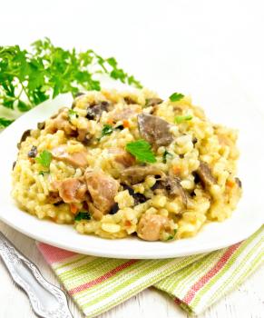 Rice risotto with mushrooms, chicken meat, cheese and garlic in a plate on a napkin, fork and parsley on the background of a light wooden board