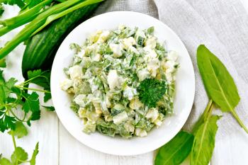 Salad of cucumber, sorrel, boiled potatoes, eggs and herbs, dressed with mayonnaise in a white plate, parsley, green onions and napkin against the background of light wooden board from above