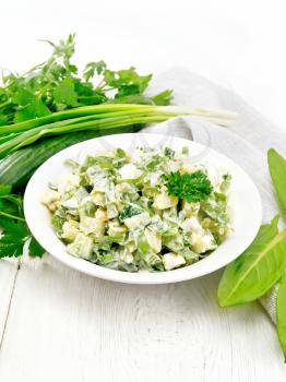 Salad of cucumber, sorrel, boiled potatoes, eggs and herbs, dressed with mayonnaise in a white plate, parsley, green onions and kitchen towel on wooden board background