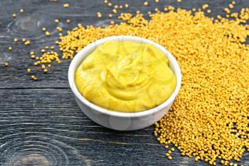 Mustard sauce in a white bowl and seeds on a wooden board background