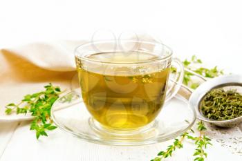 Herbal tea in a glass cup of thyme, a metal strainer with dry leaves, a linen napkin against a white wooden board