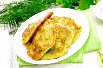 Fritters of zucchini, dill and parsley in plate on a towel, sour cream in a saucer on wooden board background