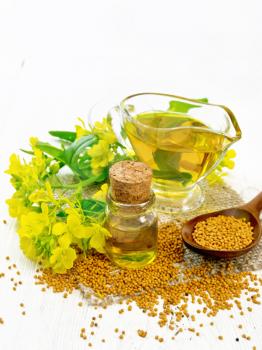 Mustard oil in a glass bottle and gravy boat, grains in a spoon and burlap, yellow mustard flowers on wooden board background