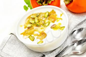 Dessert of yogurt, persimmon and honey with vanilla, cardamom and pistachios in a bowl on a towel, mint and spoons on wooden board background