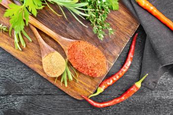 Ground hot red pepper and paprika in spoons, chili pods, spicy greens and a napkin on wooden board background from above