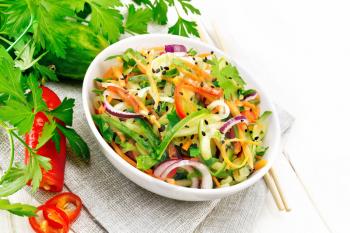 Spicy salad of cucumbers, carrots, chili peppers, purple onions, cilantro and black sesame, seasoned with vinegar and lemon juice in a bowl on a napkin against light wooden board