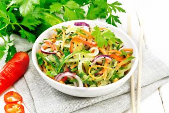 Spicy salad of cucumbers, carrots, chili peppers, purple onions, cilantro and black sesame seeds seasoned with vinegar and lemon juice in a bowl on a towel against light wooden board