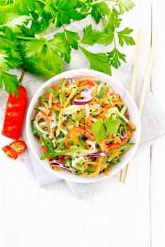 Spicy salad of cucumbers, carrots, chili peppers, purple onions, cilantro and black sesame seeds, seasoned with vinegar and lemon juice in a bowl on a napkin against light wooden board on top