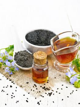 Nigella sativa oil in vial and gravy boat, seeds in a spoon and black cumin flour in a bowl on burlap, kalingi twigs with blue flowers and green leaves on light wooden board background