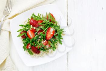 Strawberry, couscous, cedar nuts and arugula salad dressed with balsamic vinegar and olive oil in plate, a napkin and a fork on light wooden board background from above